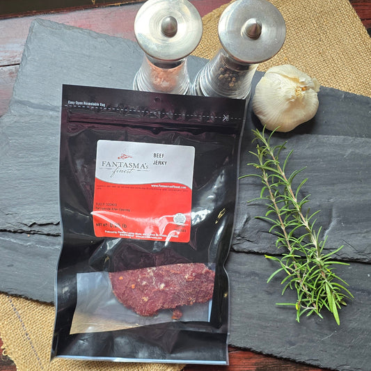 A packaged food product labeled 'Bacon' in a clear pouch with a black and red label, arranged on dark slate sheets over burlap on a rustic red wooden table, with a rosemary sprig, garlic bulb, and metal containers nearby.