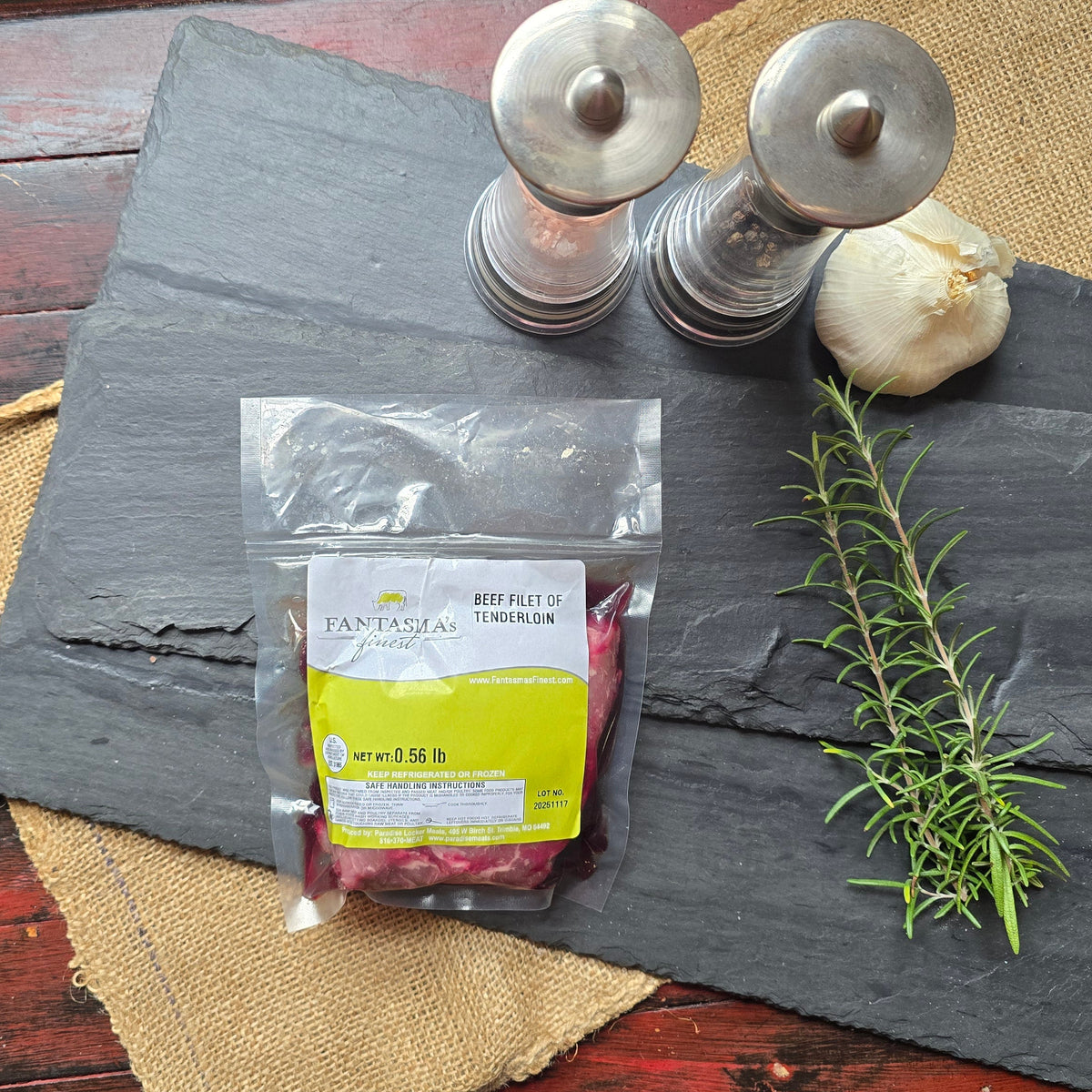 A packaged food product labeled TANKER Street Food & Drink sits on a dark slate tray, styled with a burlap runner, two metal salt and pepper mills, a garlic bulb, and a rosemary sprig on a distressed red wooden table.