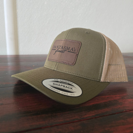An olive green and beige trucker-style snapback cap with a brown leather patch on the front reading 'FANTASMA's finest', resting on a dark wooden table against a white wall.
