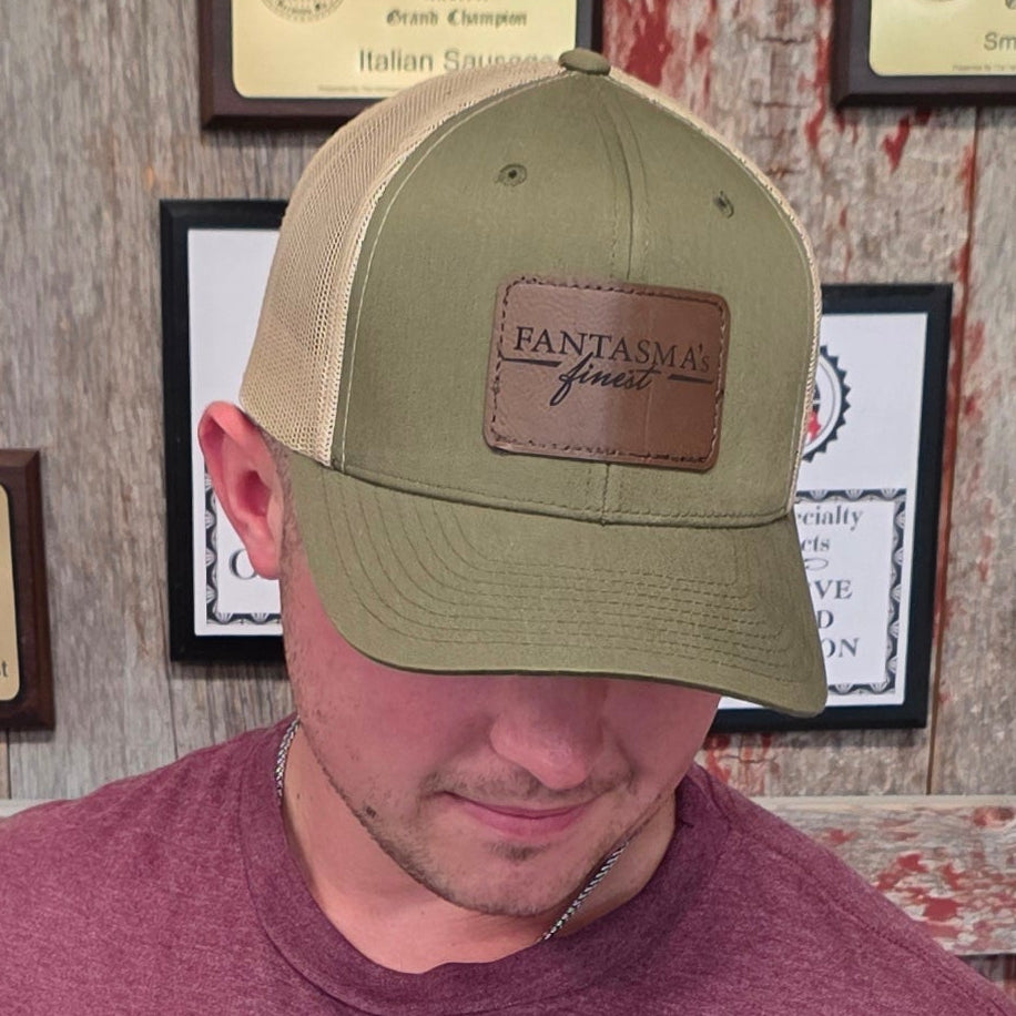 A man wearing a maroon t-shirt with white text reading 'LOCAL FARMS LOCAL BUTCHERS LOCAL TABLES' and a green trucker hat stands in front of a wall covered in framed awards and plaques.