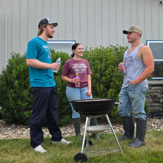 Three people standing outdoors in front of a house, gathered around a black kettle-style grill on a lawn. One person wears a bright teal t-shirt and dark pants, another wears a maroon graphic t-shirt and jeans, and the third wears a gray tank top, light blue jeans, and rubber boots.
