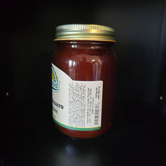 A glass jar of hot sauce with a gold metal lid and a white label, photographed on a dark black background.