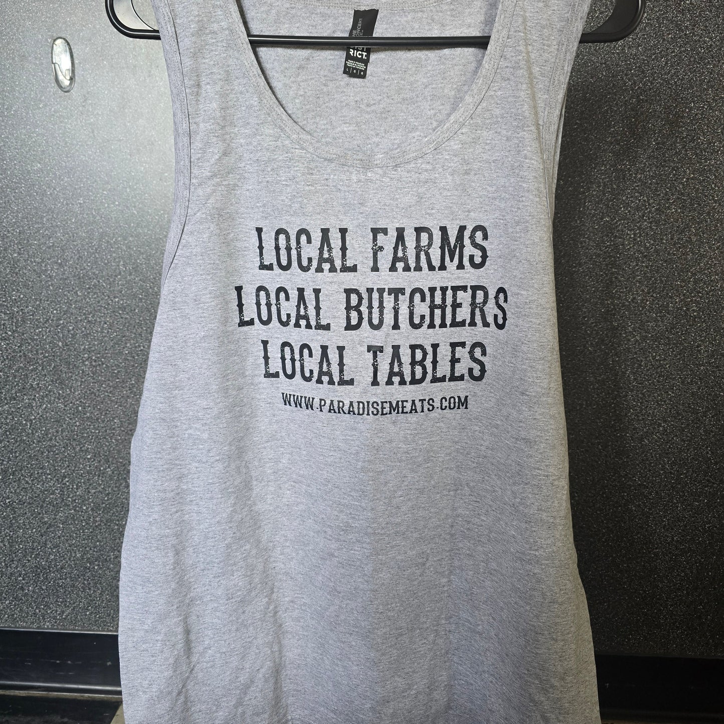 A light heather gray sleeveless tank top hanging on a black hanger, featuring the text 'LOCAL FARMS LOCAL BUTCHERS LOCAL TABLES' and a website URL printed in black on the front.