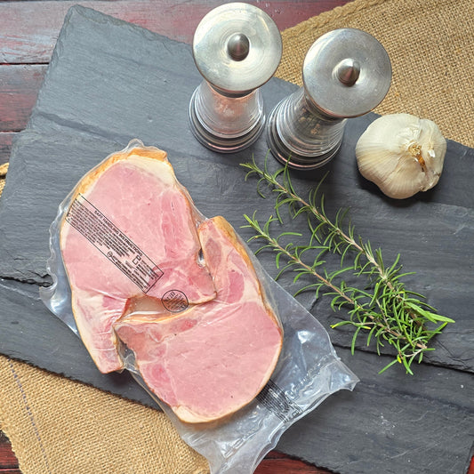 Sliced cured meat in a clear plastic wrapper on a dark slate serving board, arranged with a sprig of rosemary, a garlic bulb, and silver salt and pepper shakers on burlap and a distressed red wooden table.