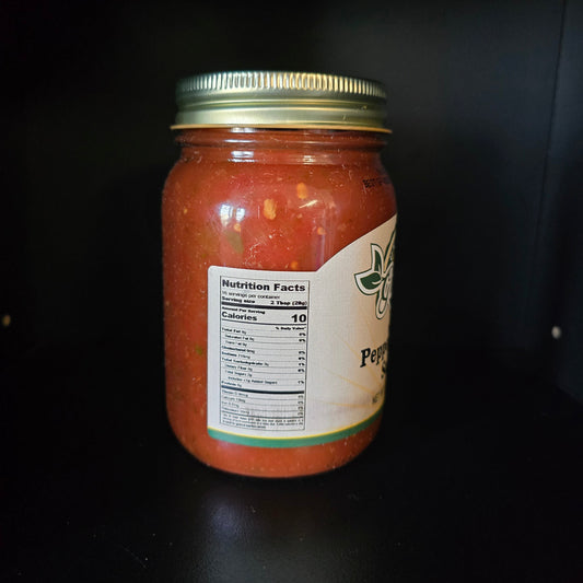 A glass jar of red pepper sauce with a gold metal lid and a partially visible cream-colored label, set against a dark black background.