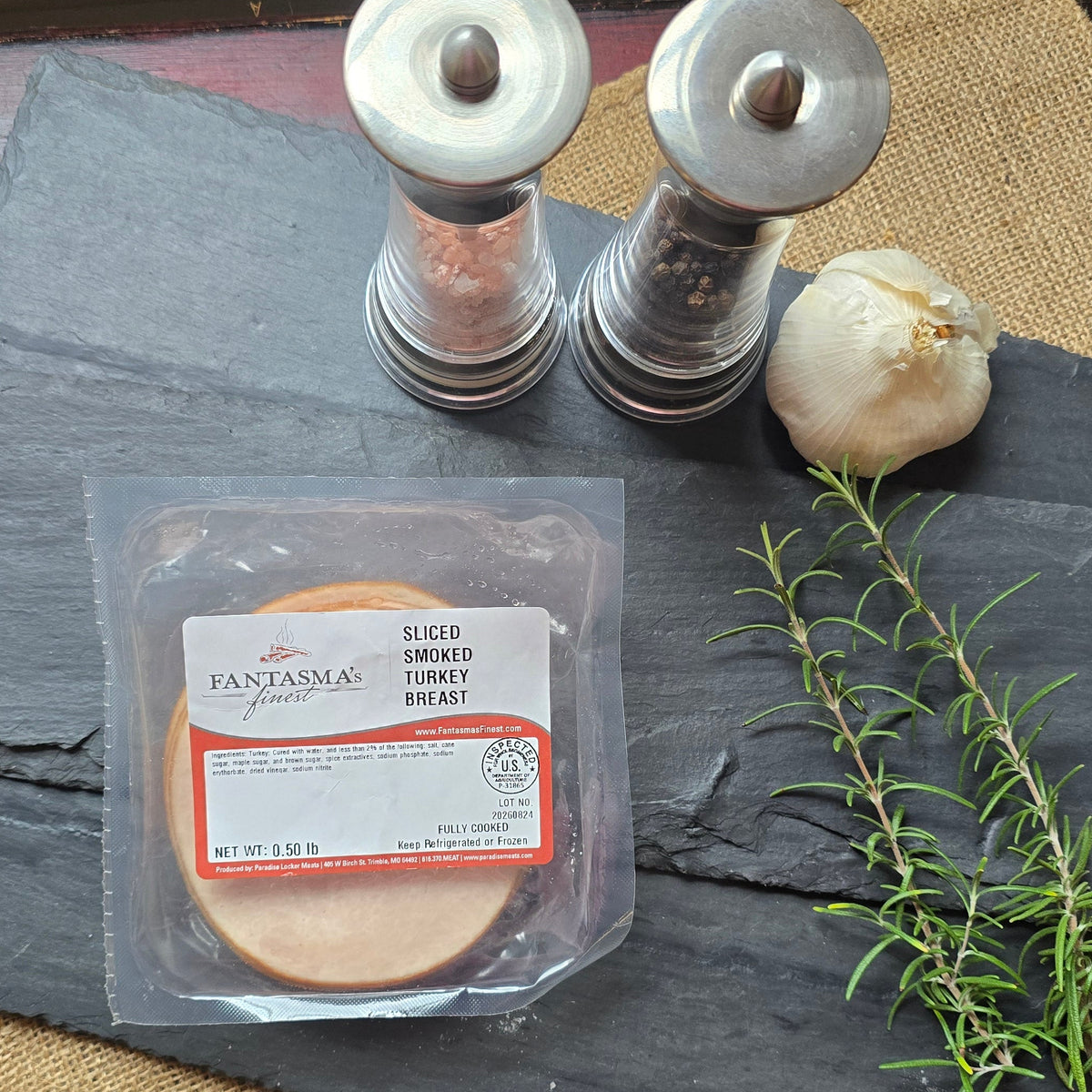 A packaged round slice of sausage or meat product sits on a dark slate board, styled with a sprig of rosemary, a garlic bulb, two salt and pepper shakers, and burlap on a distressed red wooden table.