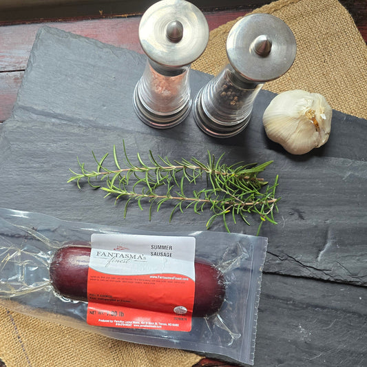 A packaged sausage log on a dark slate serving board with a sprig of rosemary, a garlic bulb, and two metal salt/pepper mills, all arranged on a rustic red wooden table with a burlap cloth.