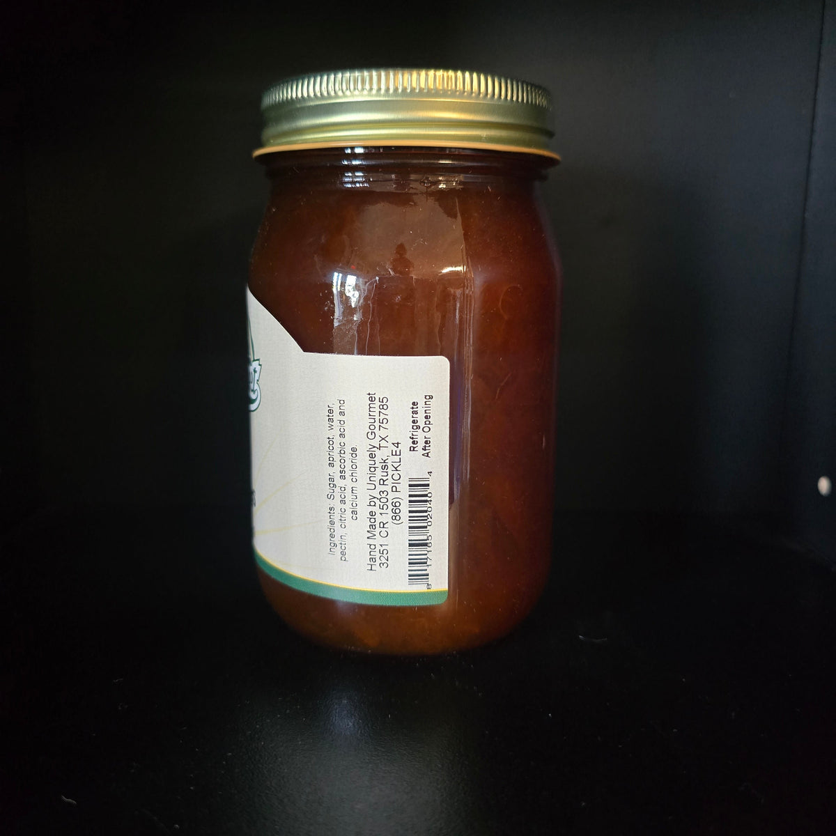 A glass jar filled with reddish-brown jam or preserve, sealed with a gold metal lid and a white label, photographed against a dark background.