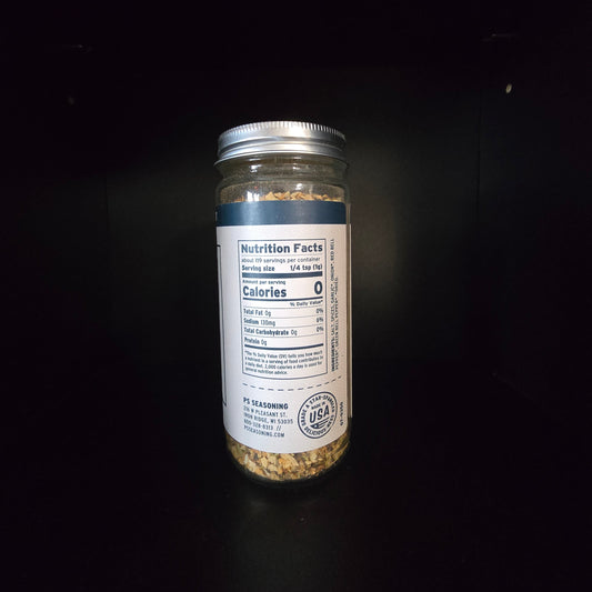 A clear glass jar filled with a beige and brown textured seasoning mix, featuring a white label with a blue stripe and a silver metal lid, set against a solid black background.