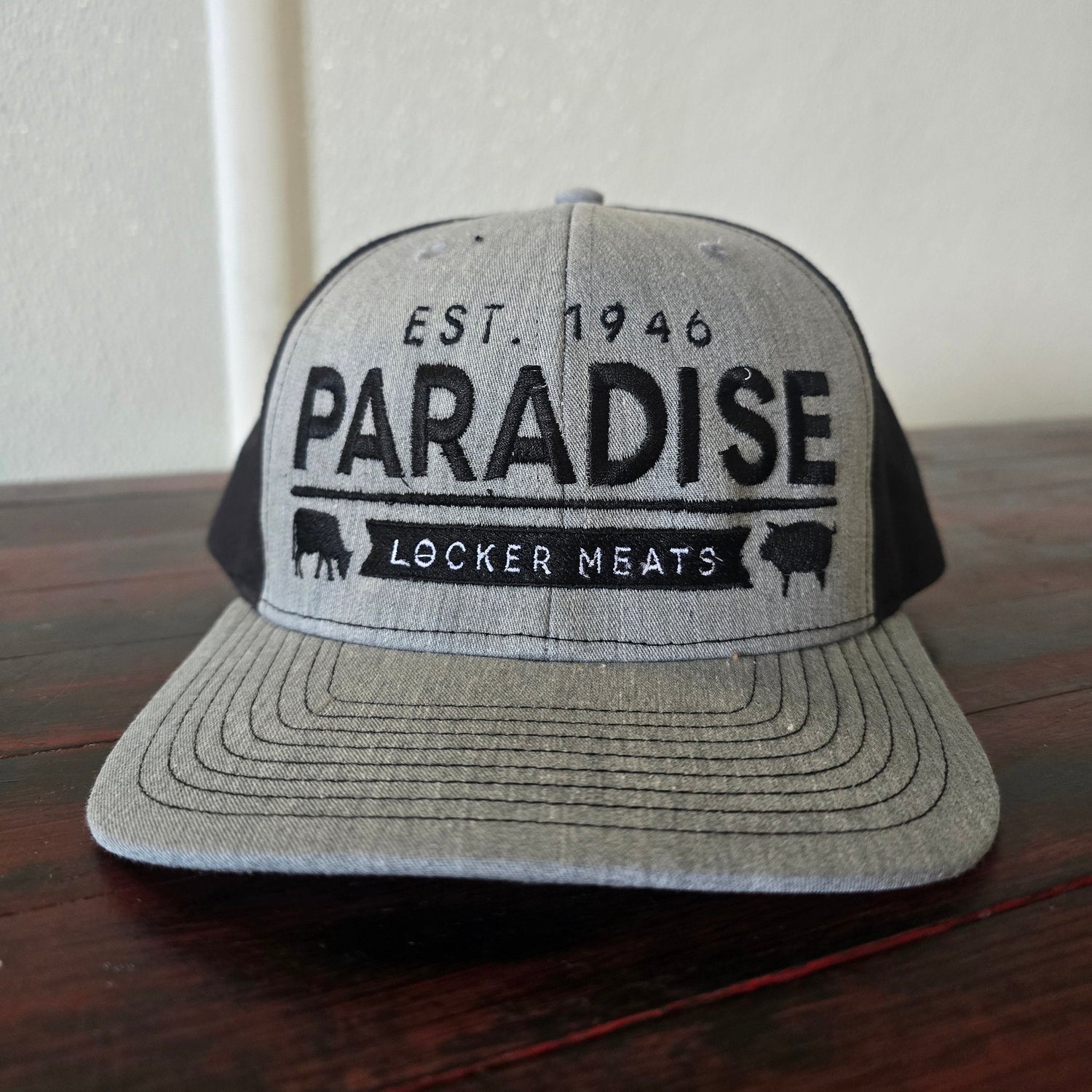 A light gray and black trucker hat with 'PARADISE LOCKER MEATS EST. 1946' printed on the front, resting on a dark wooden surface.