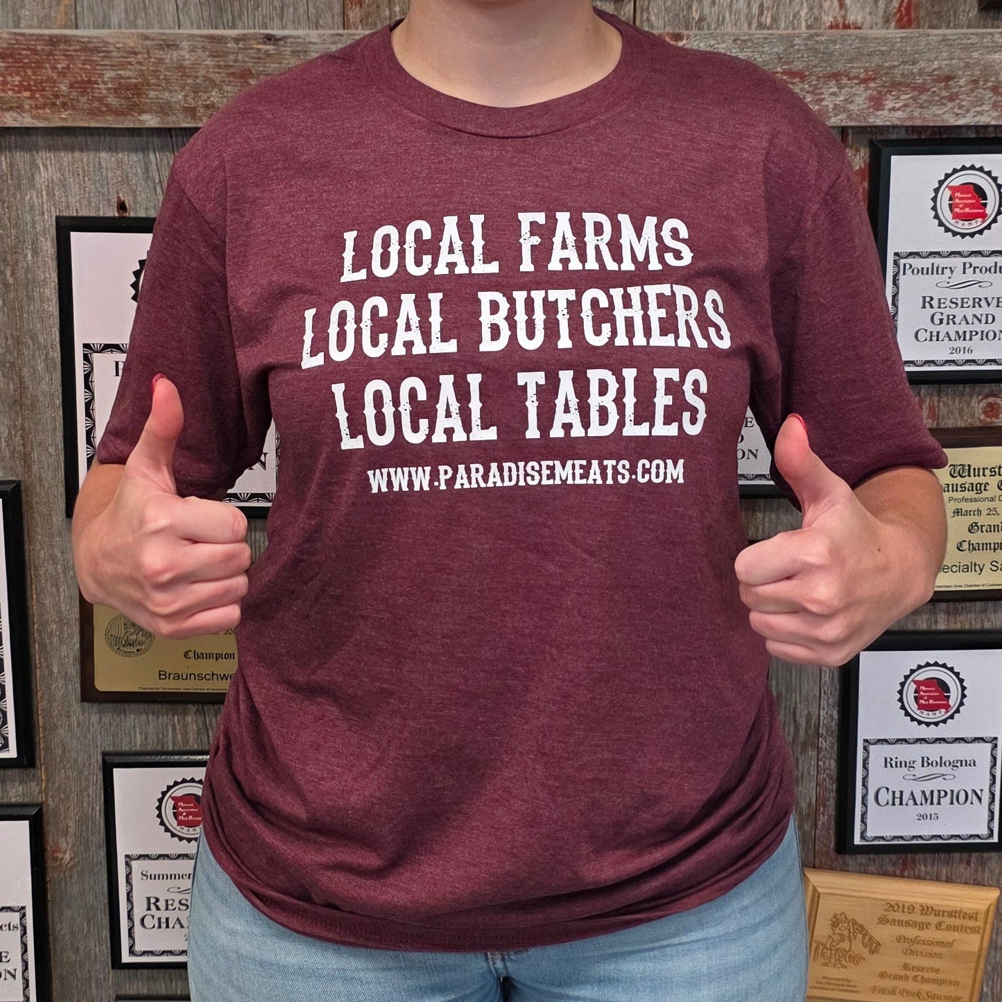A woman stands in front of a rustic wooden wall covered with framed awards and certificates, wearing a maroon graphic t-shirt that reads 'LOCAL FARMS LOCAL BUTCHERS LOCAL TABLES' with a website below, paired with light blue jeans and dark rubber boots.