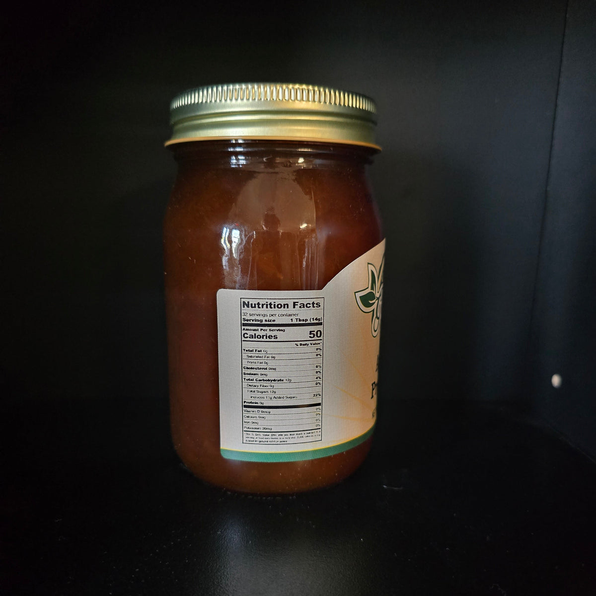 A glass jar filled with amber-brown honey, featuring a gold metal lid and a partially visible cream-colored label, set against a dark background.