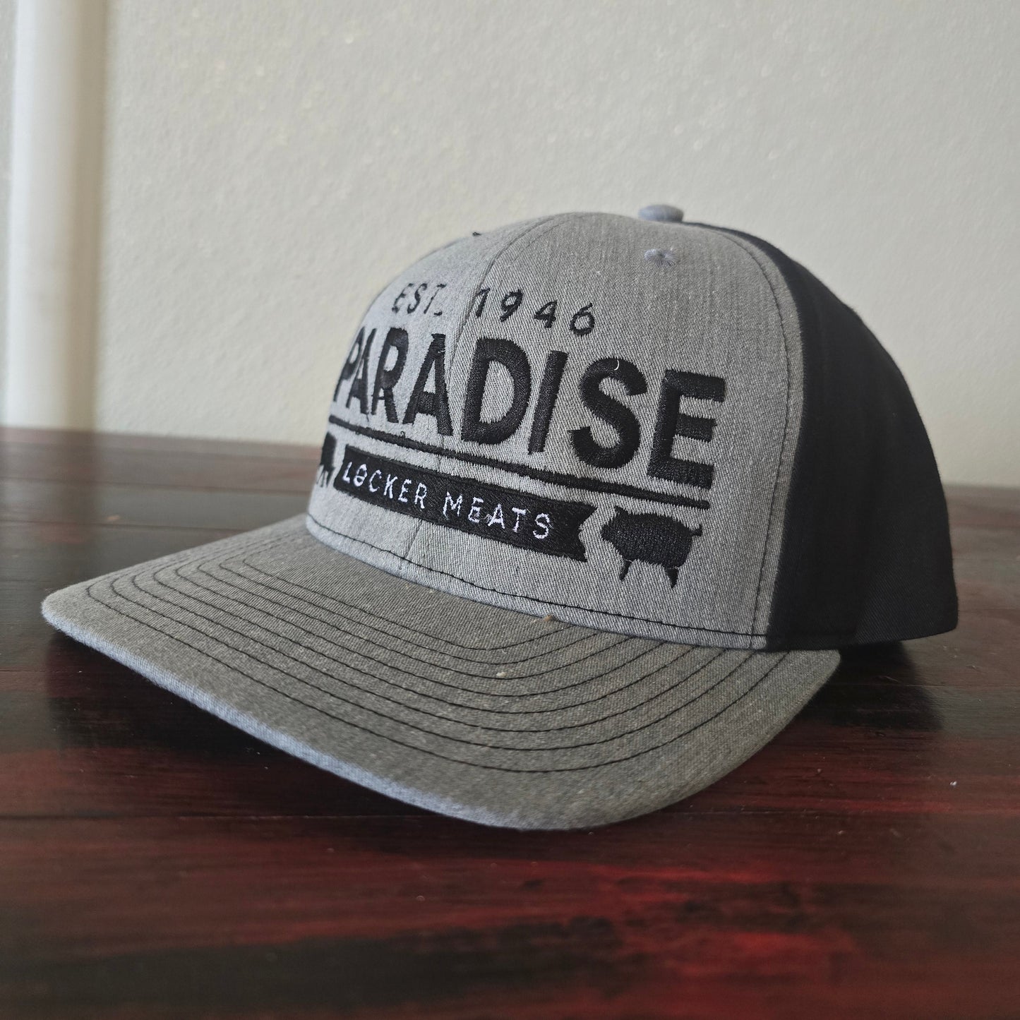 A gray and black trucker hat with 'PARADISE WACKER MEATS' printed on the front, resting on a dark wood surface against a light wall.