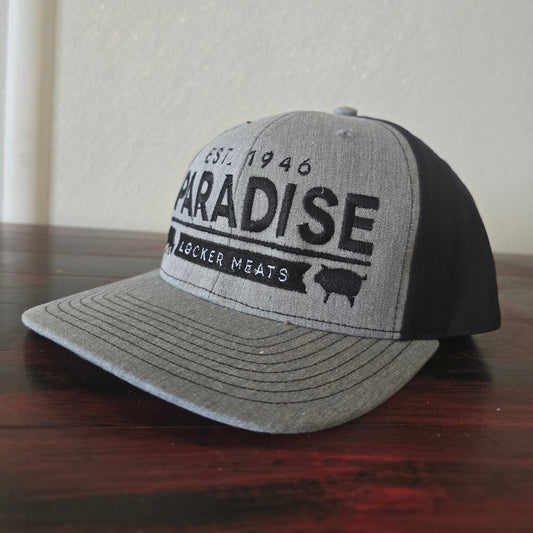 A gray and black trucker hat with 'PARADISE WACKER MEATS' printed on the front, resting on a dark wood surface against a light wall.