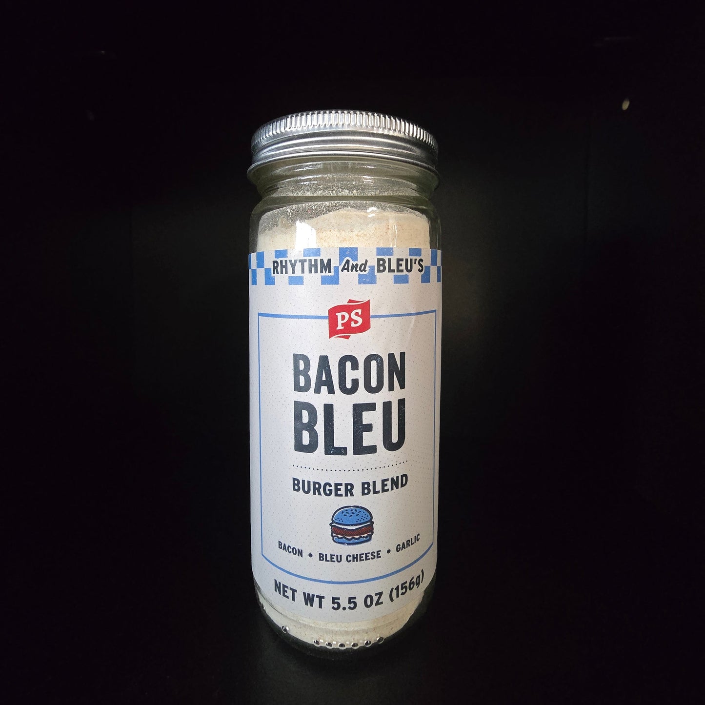 A glass jar of PS Bleu's Bacon Bleu Burger Blend seasoning, filled with a white powder, set against a solid black background.