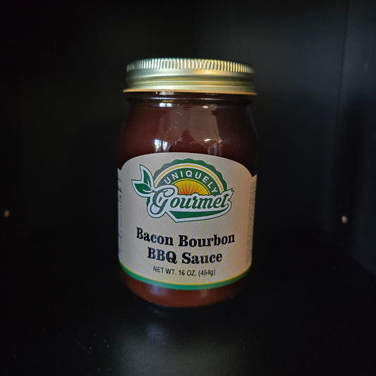 A jar of Unique Gourmet Bacon Bourbon BBQ Sauce with a gold metal lid and a white label, set against a dark background.