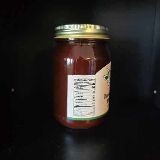 A glass jar filled with dark reddish-brown honey or syrup, featuring a gold metal lid and a partially visible cream label with green text, set against a dark black background.
