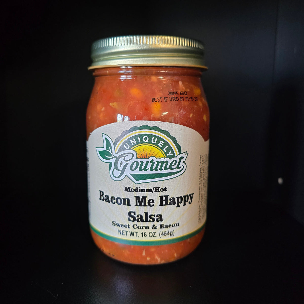 A jar of Uniquely Gourmet Bacon Me Happy Salsa, medium hot, containing orange-red salsa with visible chunks, sealed with a gold metal lid and set against a dark background.