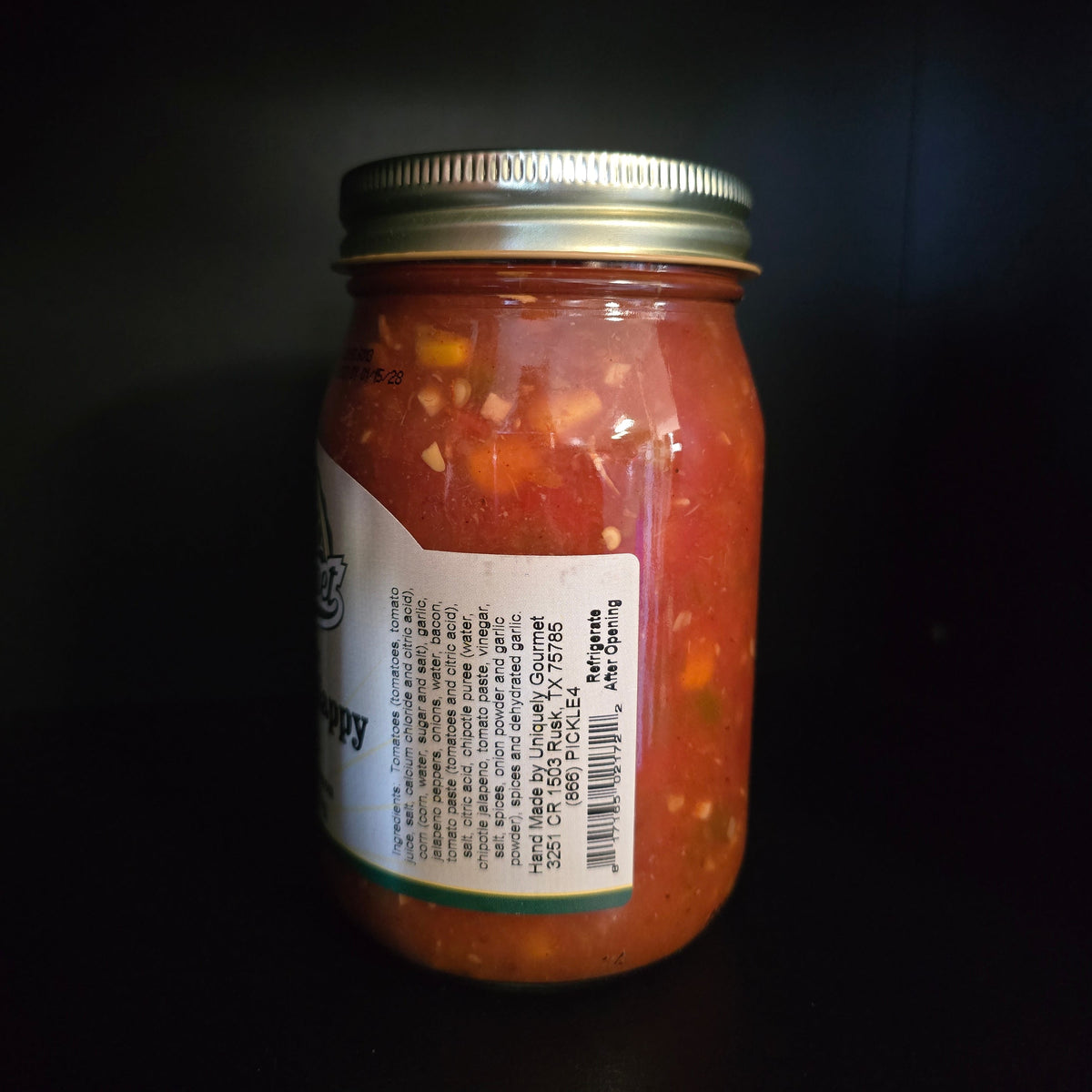 A glass jar of red salsa with a white label and a silver metal lid, photographed against a dark background.