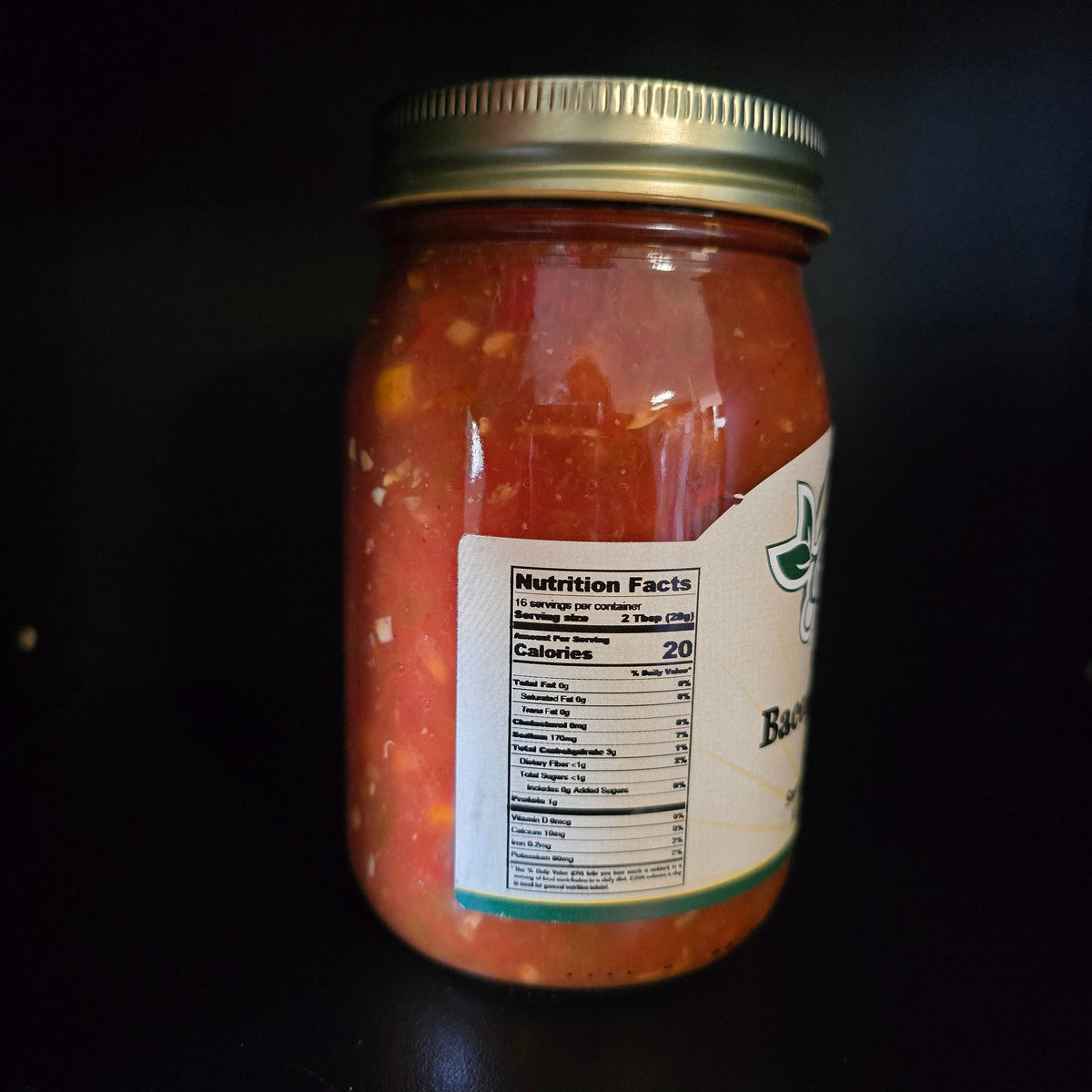 A glass jar of red salsa with a gold metal lid and a white label showing the nutrition facts, set against a dark black background.