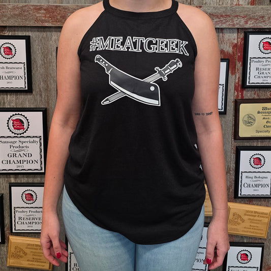 A woman stands in front of a wall covered in framed championship certificates, wearing a black halter tank top with a white graphic of a cleaver and the text '#MEATGEEK' and light blue jeans.