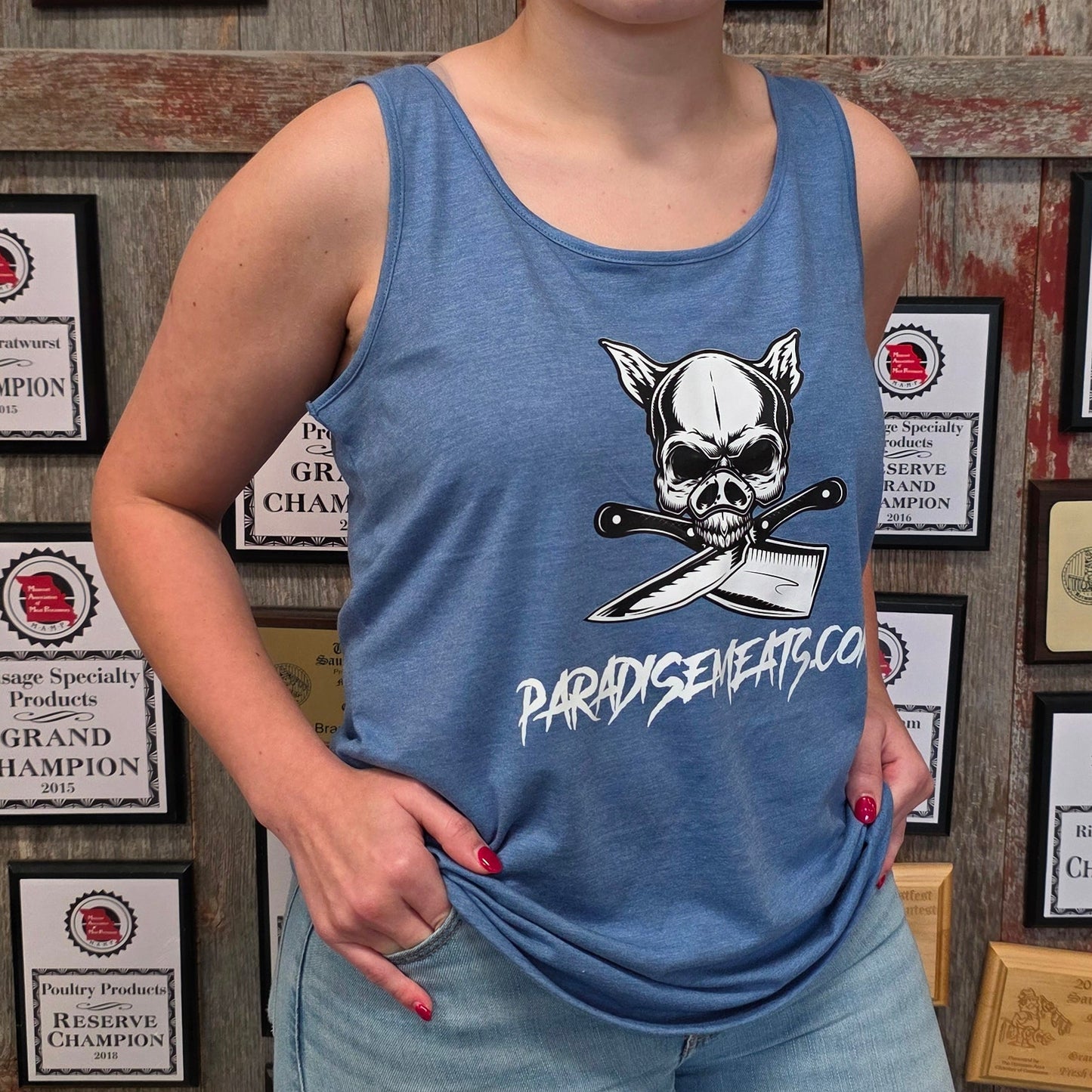 A woman wearing a blue tank top with a skull and crossbones graphic and the text 'PARDYSEVENTY' paired with light blue jeans, standing in front of a wall covered in framed championship plaques.