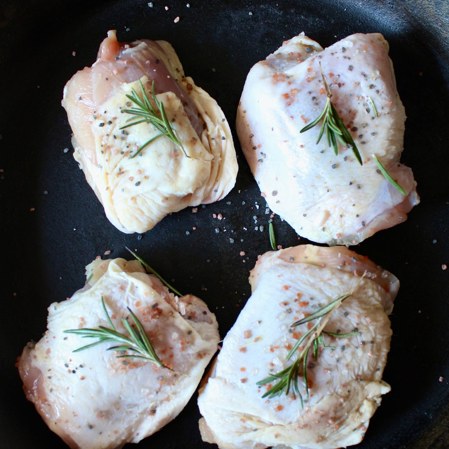 Four raw chicken thighs with rosemary on a black pan