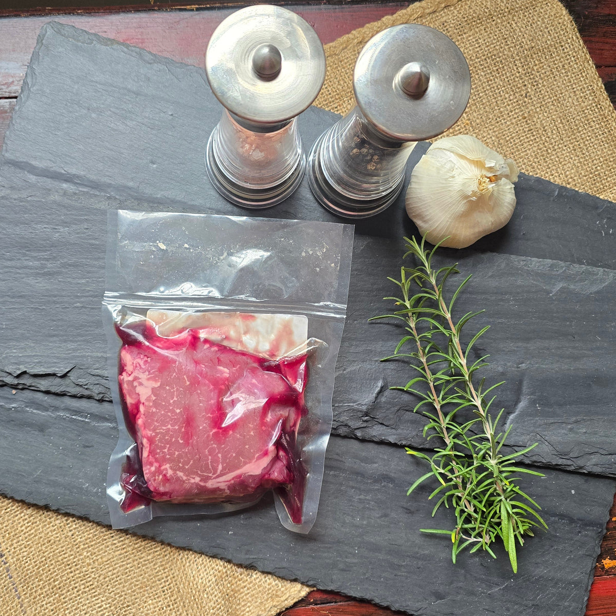 A vacuum-sealed package of raw meat sits on a dark slate board, styled with a sprig of rosemary, a whole garlic bulb, and two metal salt and pepper mills on a burlap runner over a distressed red wooden table.