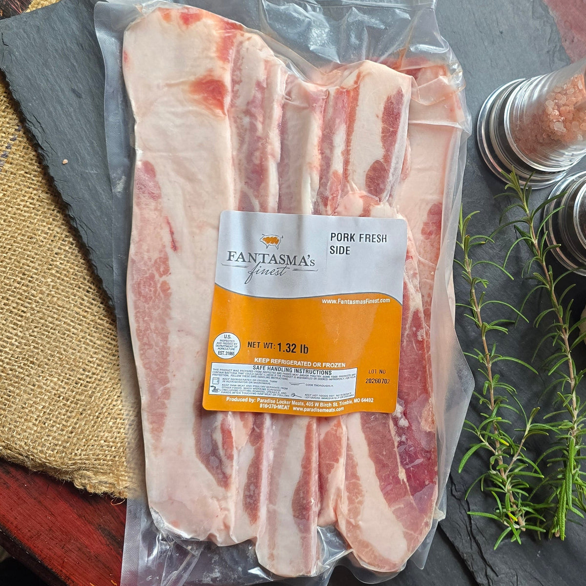 A vacuum-sealed package of Panama pork belly slices with fat trim, arranged on a rustic red wooden table with black slate boards, burlap, salt and pepper mills, rosemary, and a garlic bulb.