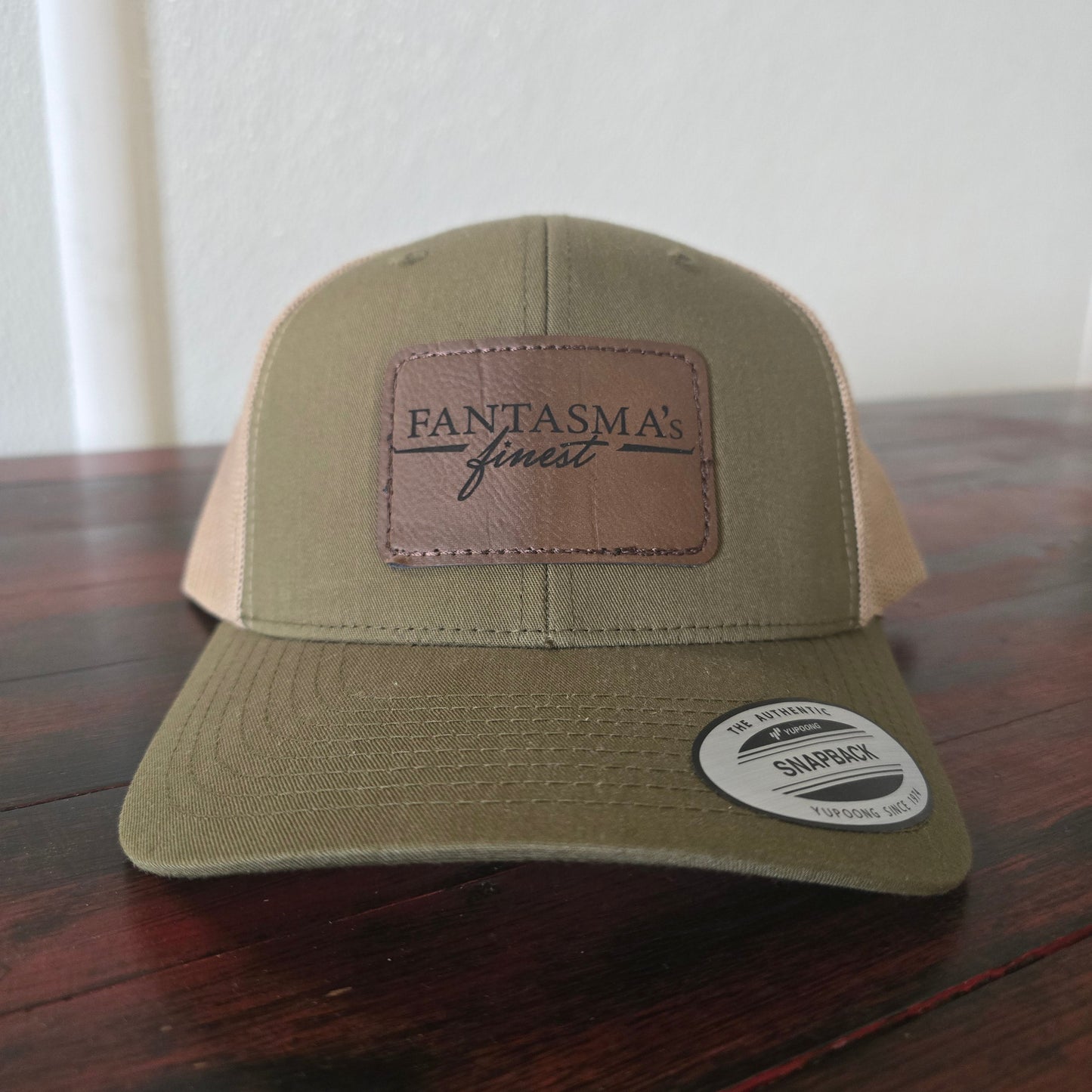 A tan and brown trucker hat with a brown leather patch on the front that reads 'FANTASMA's finest'. The hat is resting on a dark wooden table against a white wall.