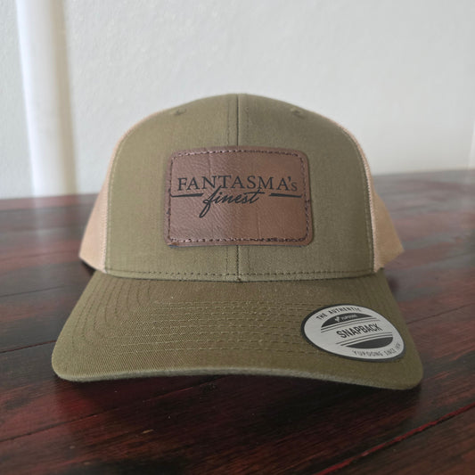 A tan and brown trucker hat with a brown leather patch on the front that reads 'FANTASMA's finest'. The hat is resting on a dark wooden table against a white wall.