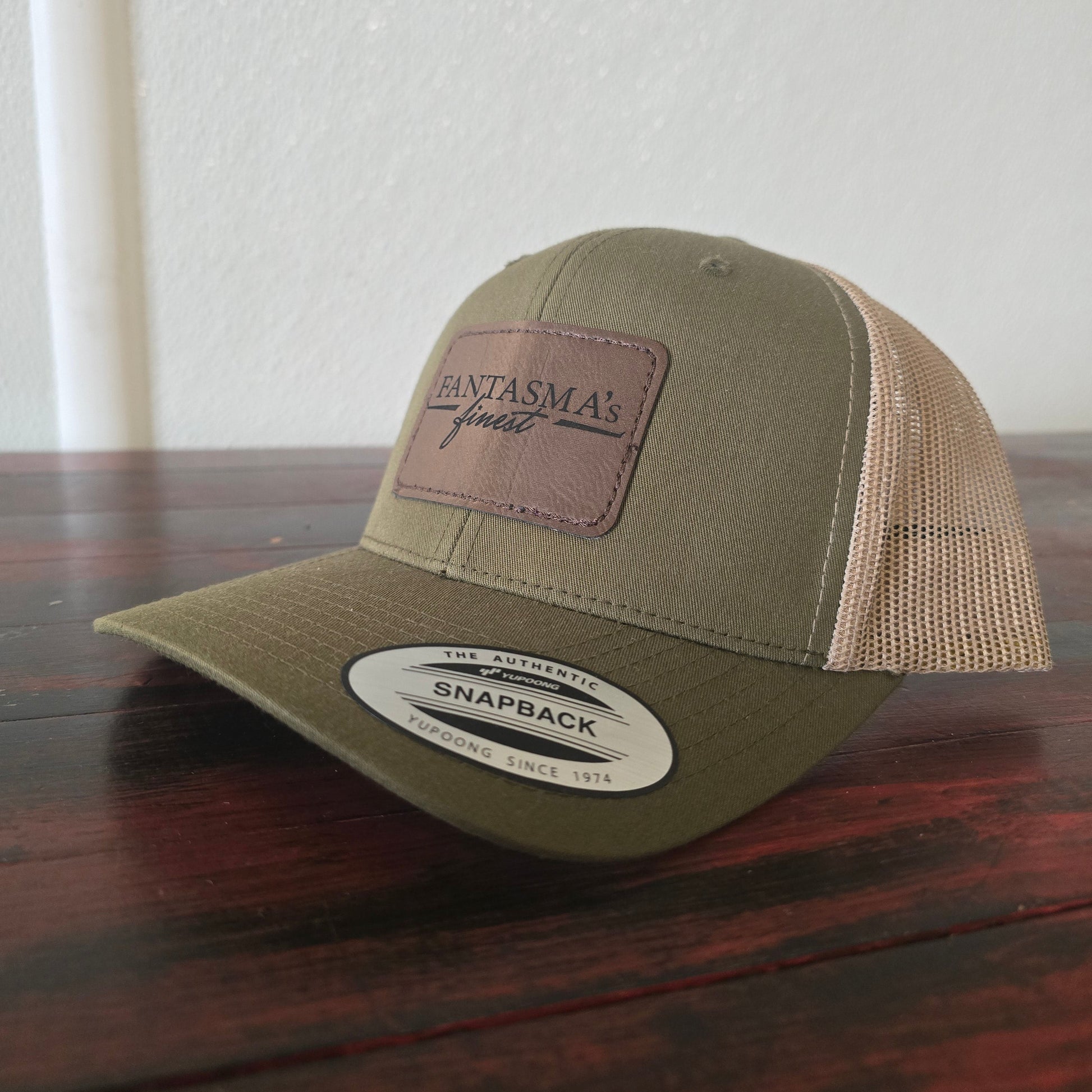 An olive green and beige trucker-style snapback cap with a brown leather patch on the front reading 'FANTASMA's finest', resting on a dark wooden table against a white wall.
