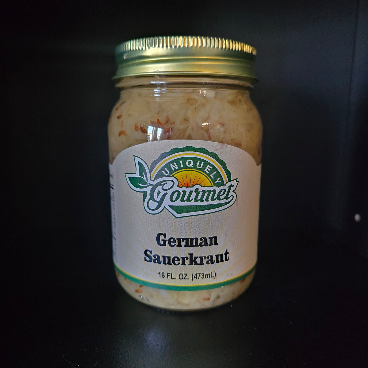 A glass jar of Gourmet German Sauerkraut with a white label and gold metal lid, set against a dark black background.
