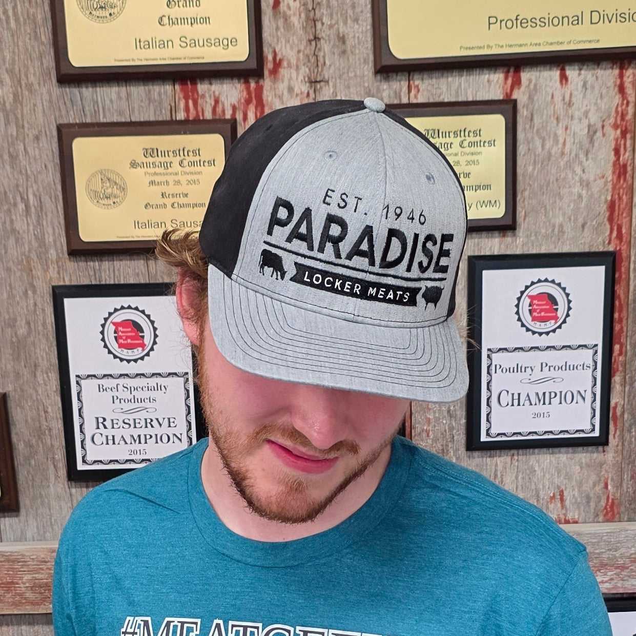 A man wearing a teal graphic t-shirt and a gray/black trucker hat stands in front of a wall covered in framed awards and certificates. The t-shirt features a blue fish graphic and the text 'SALTS & GEEKS'.