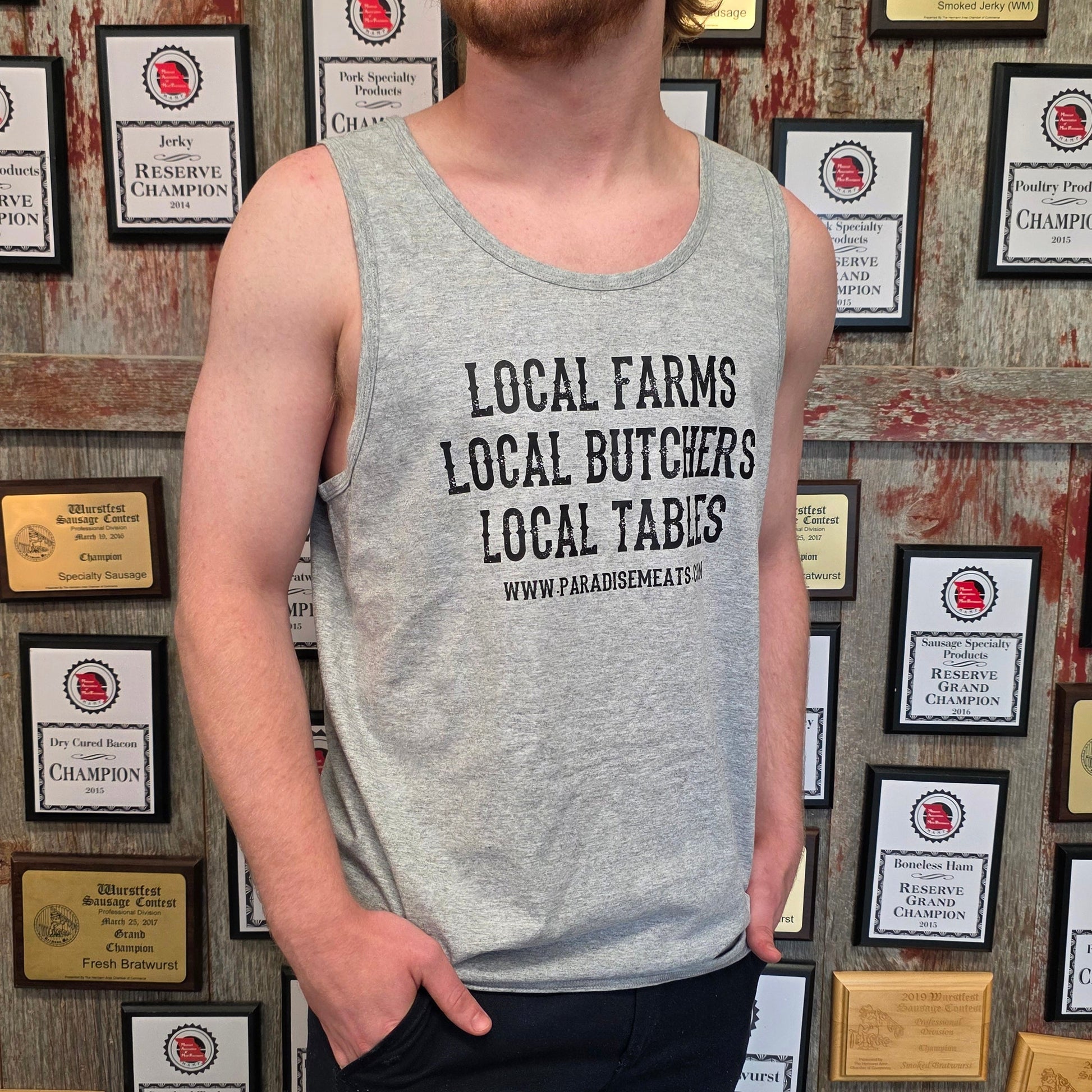 A man with red hair and a beard wearing a gray tank top with the text 'LOCAL FARMS LOCAL BUTCHERS LOCAL TABLES' stands in front of a wall covered in framed awards and plaques.
