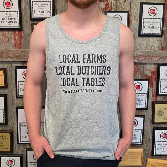 A smiling young man with red hair and a beard stands in front of a rustic wooden wall covered in framed awards and plaques. He is wearing a light gray heathered tank top with the black printed text 'LOCAL FARMS LOCAL BUTCHERS LOCAL TABLES' and a website URL, paired with dark pants.