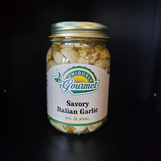 A jar of Savory Italian Garlic from uniquely Gourmet, containing sliced garlic cloves in a brine with herbs, sealed with a gold metal lid on a dark background.