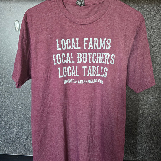 A heathered maroon short-sleeve t-shirt hanging on a wire hanger, featuring the text 'LOCAL FARMS LOCAL BUTCHERS LOCAL TABLES' in white block lettering on the front.