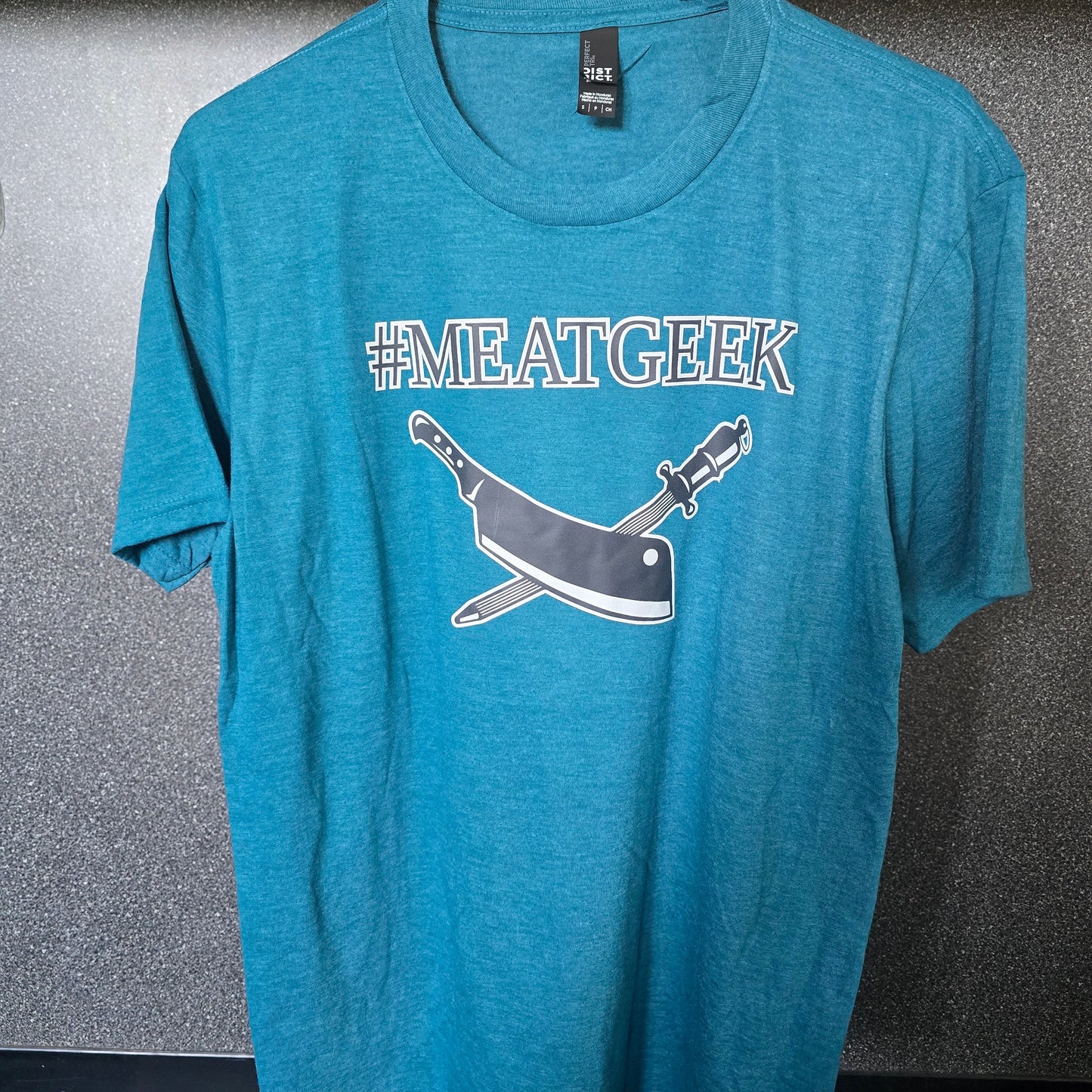 A heather teal blue t-shirt hanging on a metal hanger against a textured gray wall. The shirt features a graphic print of a cleaver and a knife crossed underneath the text '#MEATGEEK' in white and gray.