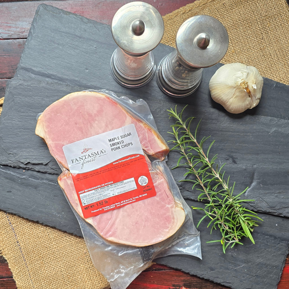 A packaged slice of ham on a dark slate board, styled with a burlap runner, salt and pepper mills, a garlic bulb, and a rosemary sprig on a distressed red wooden table.