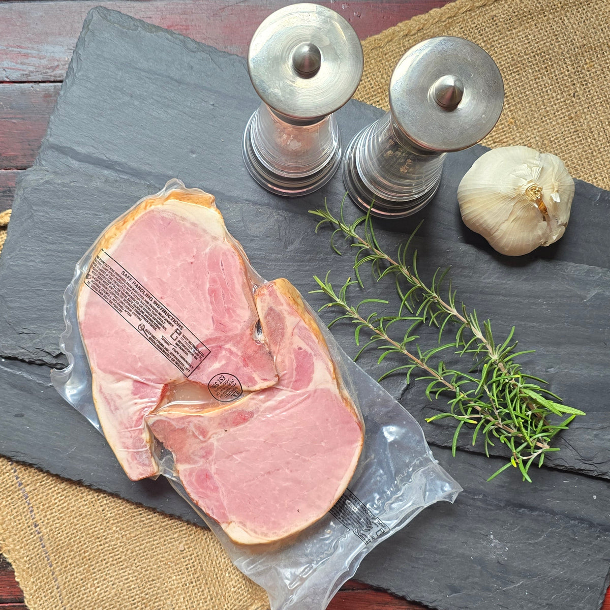 Sliced cured meat in a clear plastic wrapper on a dark slate serving board, arranged with a sprig of rosemary, a garlic bulb, and silver salt and pepper shakers on burlap and a distressed red wooden table.