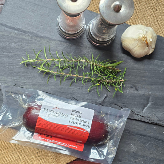 A packaged sausage stick in clear plastic packaging with a red and white label, arranged on a dark slate board with a sprig of rosemary, a garlic bulb, and two metal salt/pepper mills on a rustic red wooden table.