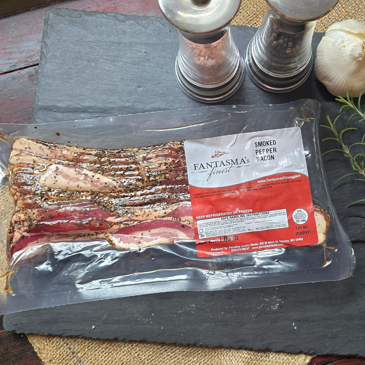 A vacuum-sealed package of smoked bacon sits on dark slate boards with a sprig of rosemary, a garlic bulb, and two metal salt/pepper mills on a rustic red wooden table.