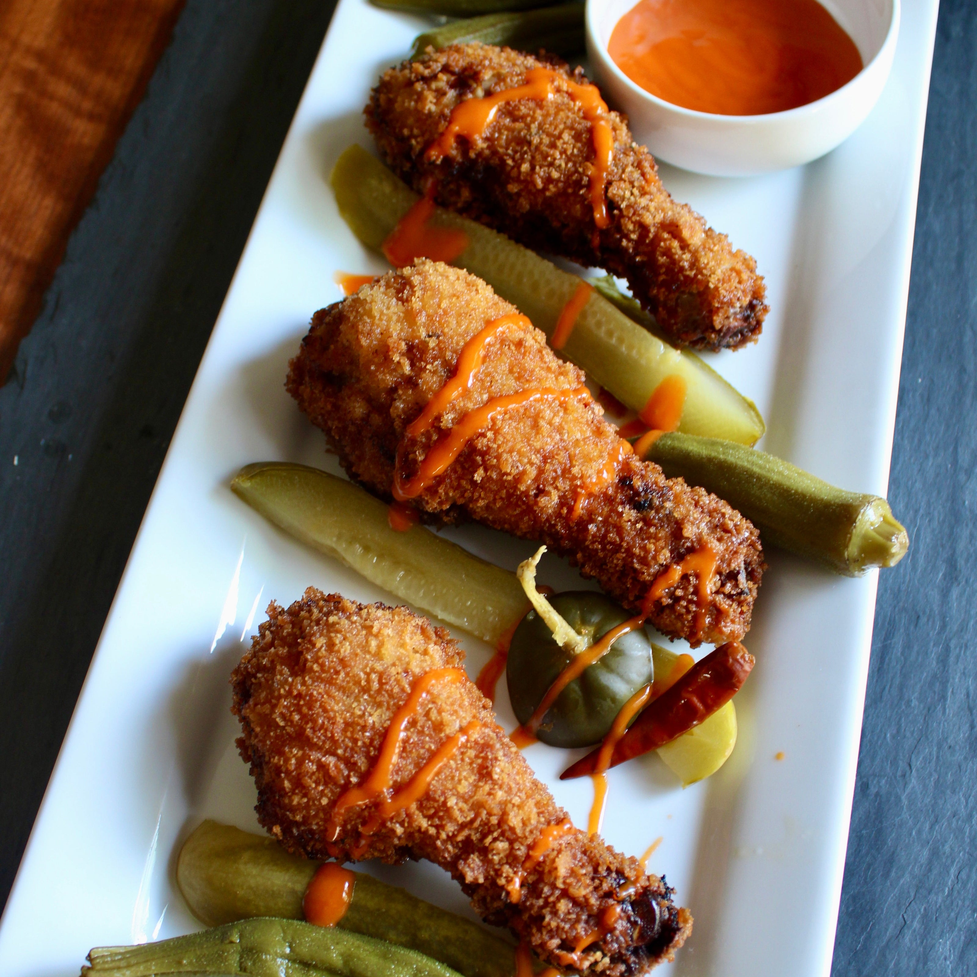 Fried chicken drumsticks with pickles and a dipping sauce on a white plate.