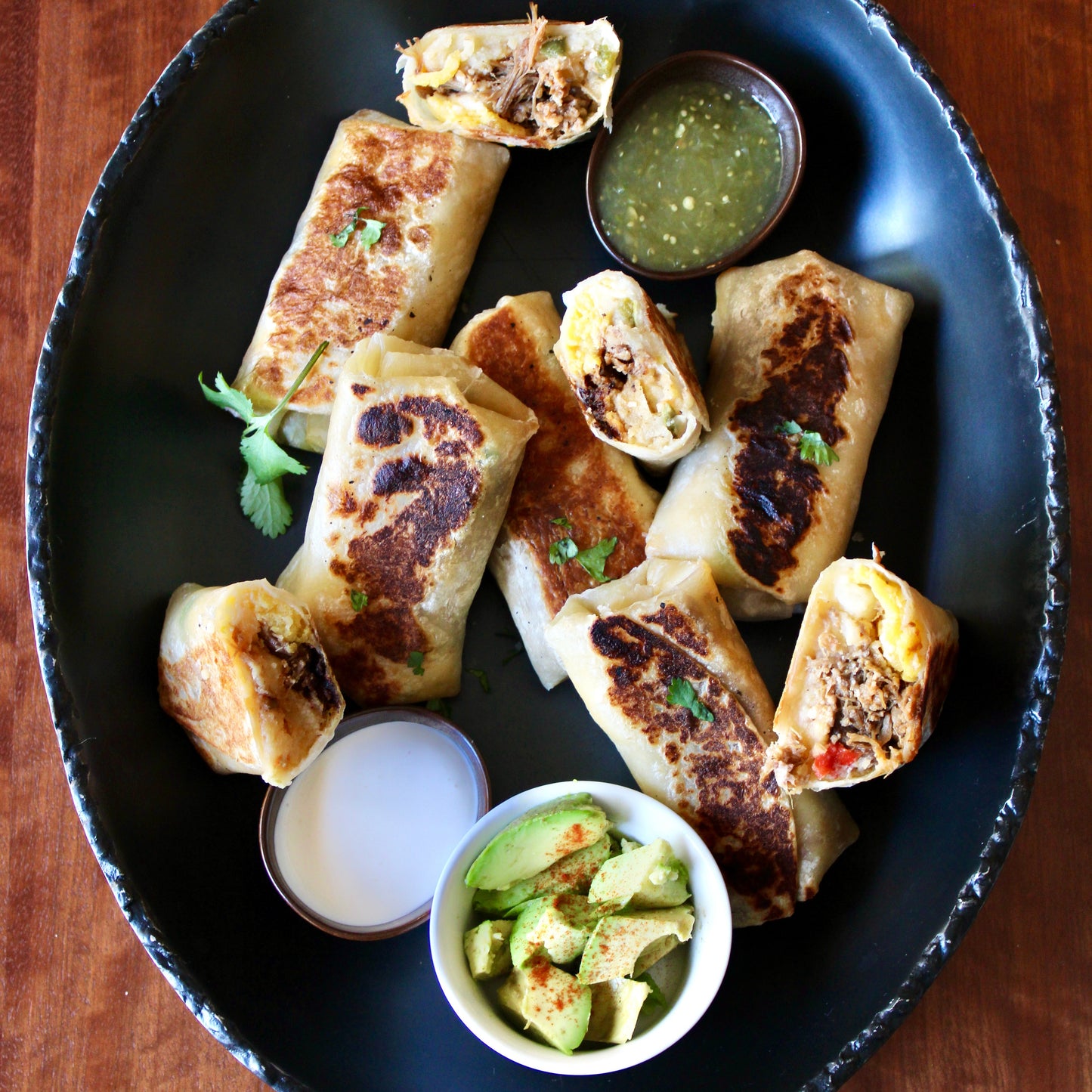 Baked burritos on a black plate with side dishes on a wooden table