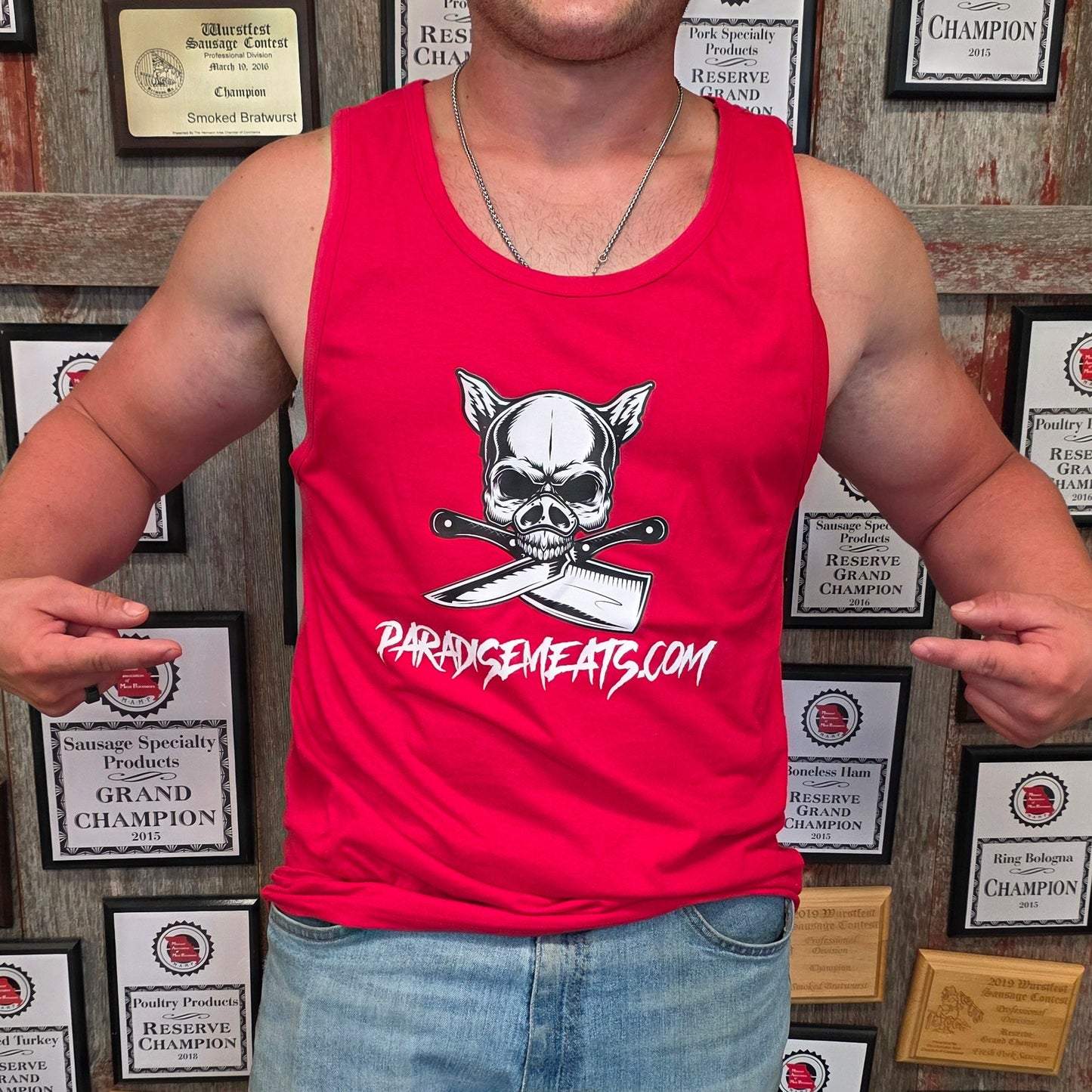 A smiling man wearing a red tank top with a skull and crossed knives graphic and the text 'PARISHMEATS.COM' stands in front of a wall covered in championship plaques.