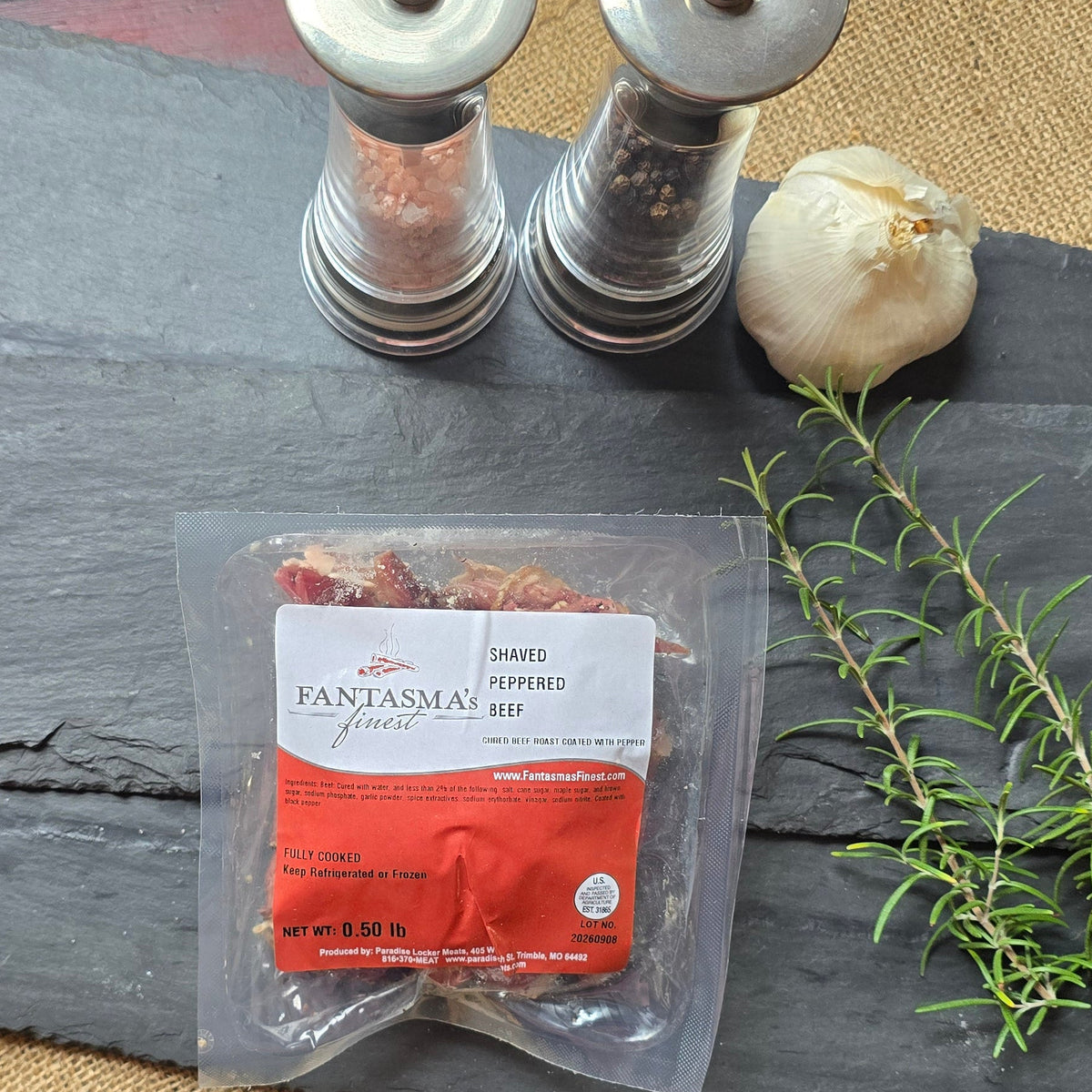 A packaged food product labeled 'PANSYONAL' sits on a dark slate board with a sprig of fresh thyme, a garlic bulb, two salt/pepper shakers, and burlap accents on a rustic red wooden table.