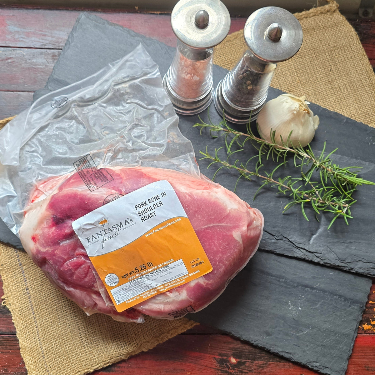 A packaged raw cut of beef placed on a rustic red wooden table, styled with a burlap runner, dark slate boards, salt and pepper mills, garlic, and rosemary. The package label shows the brand RIVIERA.