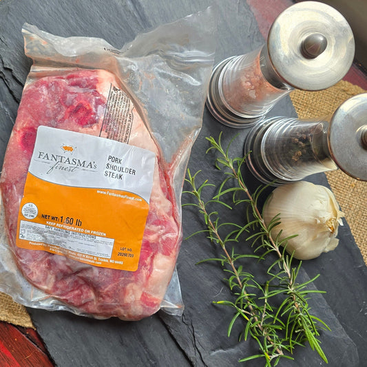 A vacuum-sealed raw cut of meat with an orange and white label sits on a dark slate board, styled with a sprig of rosemary, a garlic bulb, and two metal salt/pepper mills on a rustic red wooden table.
