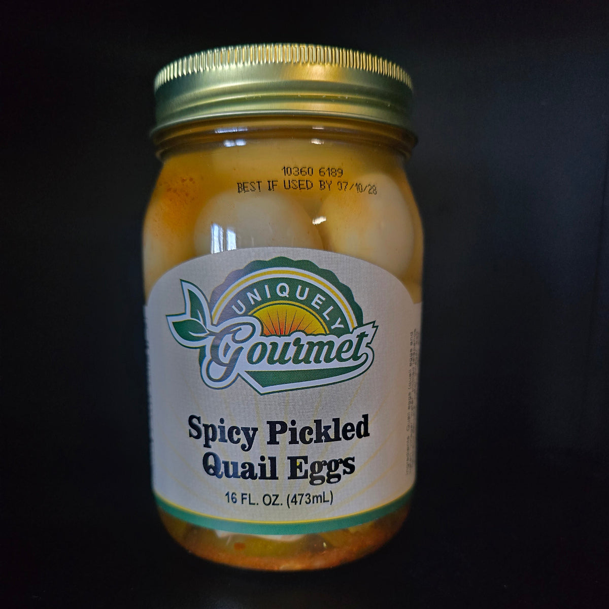 A glass jar of Unique Gourmet Spicy Pickled Quail Eggs with a gold metal lid and a white label, set against a solid black background.